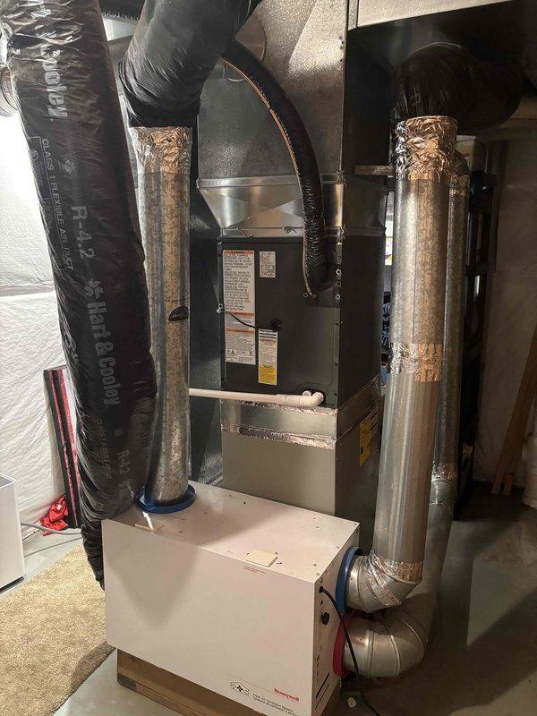 Completed full HVAC system replacement including outdoor condensing unit and indoor air handler/furnace assembly. Installed new condenser on existing concrete pad against exterior wall with proper clearances maintained. Mounted unit features horizontal louver grille design with manufacturer specifications clearly labeled. Interior installation includes new furnace unit with dual vertical metallic supply and return ductwork, properly insulated with reflective foil wrap and secured with metallic tape. Connected black flexible insulated ductwork to distribution system. Installed condensate drain pan at base of unit. Verified all copper refrigerant lines properly connected and insulated. Ensured electrical connections secure at outdoor unit. System tested and operating within manufacturer specifications.