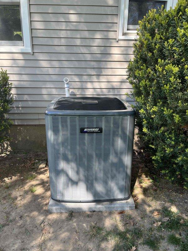 Completed full air conditioning system replacement at residential property. Removed existing outdoor condensing unit and installed new metallic gray AC condenser on existing concrete pad against exterior wall with beige vinyl siding. Unit positioned in side yard between landscaping shrubs for optimal airflow and access. Connected refrigerant lines, installed white PVC condensate drain line extending vertically from unit top, and verified all electrical connections. System tested and confirmed operational with no visible defects or irregularities.
