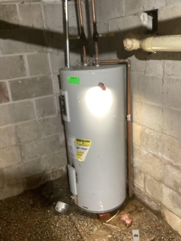 Provided estimate for replacement of existing residential water heater located in basement utility area. Unit assessed is an aging white cylindrical tank-style water heater with standard copper and metal pipe connections at top, showing visible dirt accumulation and wear consistent with extended service life. Documented current installation configuration including hot and cold water supply lines, electrical connections via wall-mounted conduit box, and proper venting system with PVC drainage components for customer's consideration of replacement options.