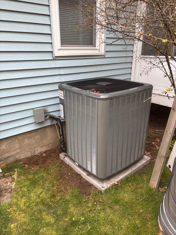 Completed full air conditioning system replacement including outdoor condensing unit and indoor air handler components. New gray outdoor AC unit installed on concrete pad with proper refrigerant line connections and electrical hookup, positioned against exterior wall with appropriate clearances. Indoor Carrier system integrated with existing ductwork infrastructure, including proper PVC condensate drain line installation and ventilation connections to ensure optimal system performance and code compliance.