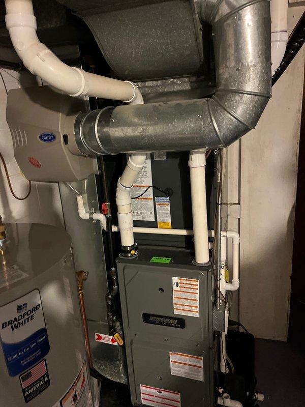 Completed full air conditioning system replacement including outdoor condensing unit and indoor air handler components. New gray outdoor AC unit installed on concrete pad with proper refrigerant line connections and electrical hookup, positioned against exterior wall with appropriate clearances. Indoor Carrier system integrated with existing ductwork infrastructure, including proper PVC condensate drain line installation and ventilation connections to ensure optimal system performance and code compliance.