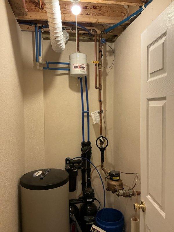 Responded to emergency plumbing leak in utility room ceiling where water line was dripping into electrical breaker box when water supply was activated. Upon arrival, found customer had shut off main water supply as a safety precaution. Inspected utility room with exposed ceiling joists, PEX piping system, water treatment equipment, and electrical panel. Assessed leak source in overhead water supply line and potential electrical hazards from water intrusion into breaker panel. Documented conditions of plumbing infrastructure including blue PEX lines, copper piping, water softener system, and exterior backflow preventer assembly. Coordinated with customer regarding necessary repairs to resolve leak and restore safe water service to the residence.