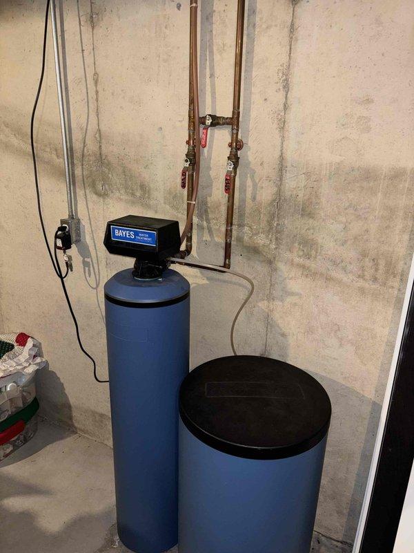 Responded to plumbing lead for leaking water softener system. Upon inspection, identified leak originating from water softener installation consisting of dual-tank Bayes system with tall brine tank and shorter resin tank. Examined copper supply piping with red-handled shut-off valves and connections to treatment system. Assessed aging copper piping showing corrosion and patina, along with newer PVC drainage components. Documented existing plumbing configuration including pump unit and electrical connections for water treatment system. Photographed installation to record current condition of equipment, piping connections, and potential leak sources for repair recommendations.