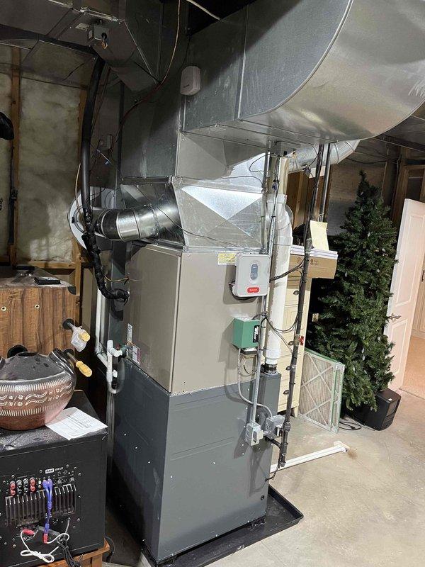 Completed full furnace installation in residential garage/utility space. Installed new gray vertical furnace unit with proper mounting and positioning on protective base mat. Connected metallic supply and return ductwork with curved elbow joints and cylindrical sections, secured with metal straps and brackets to ceiling. Installed white PVC intake and exhaust venting system extending from furnace unit. Mounted and wired electrical control panel and junction boxes to wall per code requirements. Verified all ductwork connections are properly sealed and secured. Tested system operation, confirmed proper airflow, and ensured all electrical connections are functioning correctly. Installation complete and system operational.