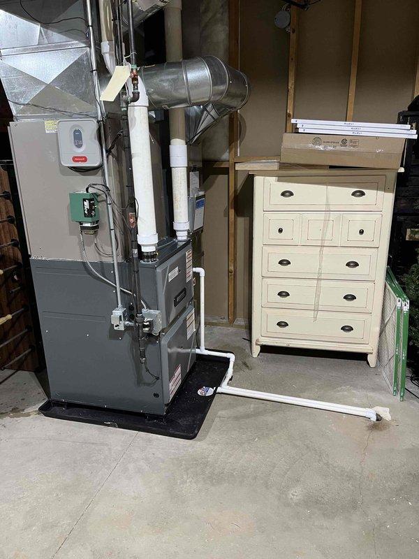 Completed full furnace installation in residential garage/utility space. Installed new gray vertical furnace unit with proper mounting and positioning on protective base mat. Connected metallic supply and return ductwork with curved elbow joints and cylindrical sections, secured with metal straps and brackets to ceiling. Installed white PVC intake and exhaust venting system extending from furnace unit. Mounted and wired electrical control panel and junction boxes to wall per code requirements. Verified all ductwork connections are properly sealed and secured. Tested system operation, confirmed proper airflow, and ensured all electrical connections are functioning correctly. Installation complete and system operational.