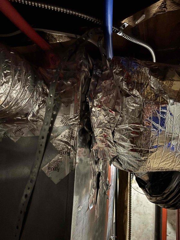 Responded to complaint of inadequate heating and cooling to upstairs area on recently installed EnergiAir system. Upon inspection of attic space, discovered extensive damage to flexible ductwork serving the upper level. The outer foil vapor barrier layer was severely torn and shredded in multiple locations, with insulation exposed and hanging loose from the main duct body. This compromised ductwork was allowing significant conditioned air loss before reaching the intended living spaces, resulting in poor temperature control upstairs despite the system operating normally. Documented the damaged ductwork conditions with photographs showing the deteriorated sections and compromised insulation integrity. The torn and peeling metallic outer layer indicated either installation damage or subsequent physical impact to the duct system. Recommended immediate ductwork replacement or repair to restore proper airflow delivery and system performance to the upstairs zones.