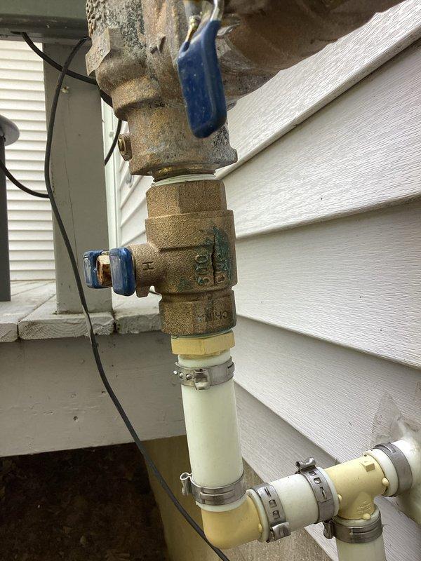 Responded to plumbing leak emergency at residential property approximately 25 years old. Upon arrival, identified cracked and leaking water pump with leak confined to immediate pump area. Inspected exterior plumbing assembly mounted on building foundation, which included brass valve fittings, backflow preventer, blue-handled shut-off valves, and PVC piping connections. Noted significant corrosion and weathering on brass components consistent with age of home. Assessed vertical pipe assembly and connection points between metal and plastic piping sections secured with hose clamps. Documented condition of all plumbing components including valve positions, pipe transitions, and irrigation system controls. Photographed entire installation showing corroded brass fittings, white PVC pipes, elbow connections, and associated electrical wiring. Leak was successfully contained and system evaluated for proper functionality. Recommended monitoring of corroded brass components for potential future r