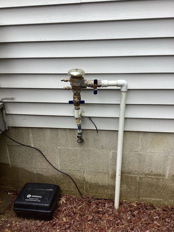 Responded to plumbing leak emergency at residential property approximately 25 years old. Upon arrival, identified cracked and leaking water pump with leak confined to immediate pump area. Inspected exterior plumbing assembly mounted on building foundation, which included brass valve fittings, backflow preventer, blue-handled shut-off valves, and PVC piping connections. Noted significant corrosion and weathering on brass components consistent with age of home. Assessed vertical pipe assembly and connection points between metal and plastic piping sections secured with hose clamps. Documented condition of all plumbing components including valve positions, pipe transitions, and irrigation system controls. Photographed entire installation showing corroded brass fittings, white PVC pipes, elbow connections, and associated electrical wiring. Leak was successfully contained and system evaluated for proper functionality. Recommended monitoring of corroded brass components for potential future r