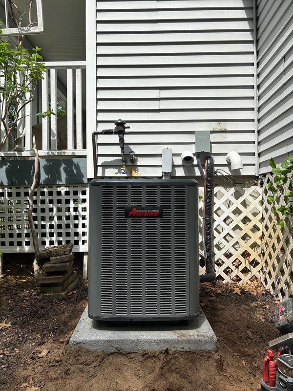 Completed full air conditioning system replacement at residential property. Removed existing outdoor condenser unit and installed new dark gray HVAC condenser on concrete pad in backyard location adjacent to house exterior. Positioned unit on stable concrete foundation with proper clearances from white vinyl siding and lattice skirting. Ran and secured refrigerant lines and electrical conduit from condenser unit up exterior wall to connect with indoor air handler components. All copper line sets properly insulated and fastened to building structure per manufacturer specifications. System installed with appropriate clearances from decorative lattice panels and landscaping features to ensure adequate airflow and serviceability. Unit positioned to minimize visual impact while maintaining accessibility for future maintenance. Installation completed with all refrigerant connections brazed, pressure tested, and evacuated prior to charging system. Electrical connections made to outdoor discon