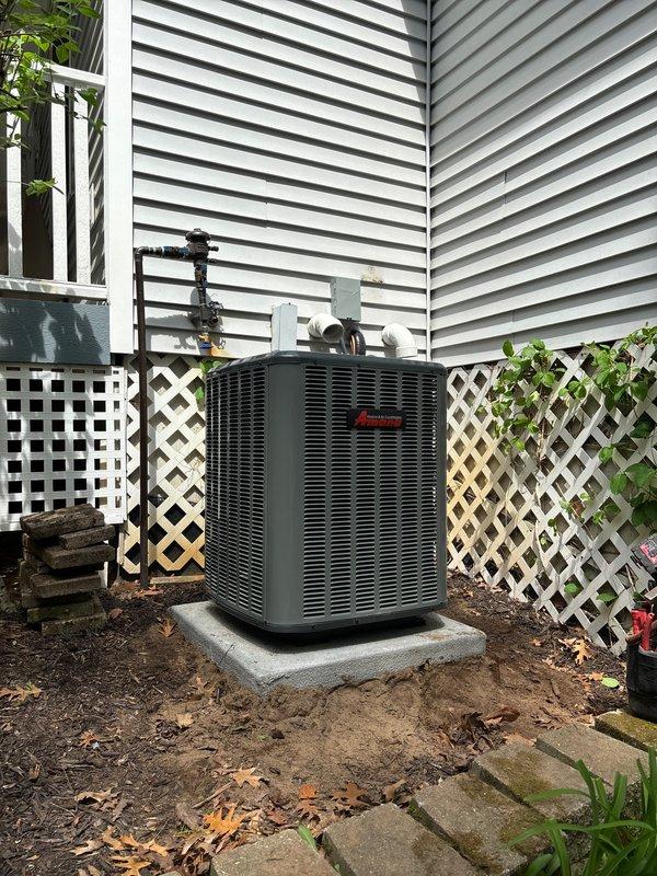 Completed full air conditioning system replacement at residential property. Removed existing outdoor condenser unit and installed new dark gray HVAC condenser on concrete pad in backyard location adjacent to house exterior. Positioned unit on stable concrete foundation with proper clearances from white vinyl siding and lattice skirting. Ran and secured refrigerant lines and electrical conduit from condenser unit up exterior wall to connect with indoor air handler components. All copper line sets properly insulated and fastened to building structure per manufacturer specifications. System installed with appropriate clearances from decorative lattice panels and landscaping features to ensure adequate airflow and serviceability. Unit positioned to minimize visual impact while maintaining accessibility for future maintenance. Installation completed with all refrigerant connections brazed, pressure tested, and evacuated prior to charging system. Electrical connections made to outdoor discon