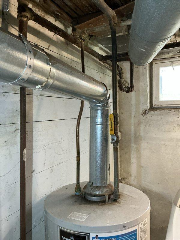 Completed on-site assessment of existing water heater installation in basement utility area. Documented current unit specifications, venting configuration with metallic exhaust ductwork, plumbing connections including shut-off valves and piping, and overall installation conditions. Photographed equipment labels, surrounding infrastructure, and workspace layout to provide comprehensive estimate for water heater replacement.