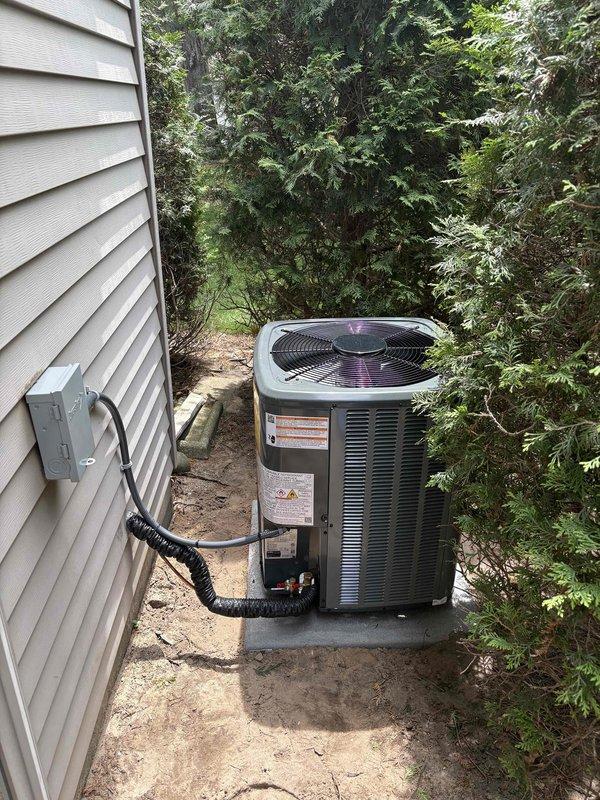 Completed full installation of new furnace and air conditioning system at residential property. Outdoor condensing unit installed on concrete pad against exterior wall with proper clearances for airflow and maintenance access. Electrical disconnect box mounted to building with flexible conduit providing power connection to unit. Refrigerant lines, copper piping, and brass fittings connected and secured at condensing unit base. All manufacturer labels, wiring diagrams, and safety placards properly affixed to equipment. System installed per manufacturer specifications and local code requirements with appropriate clearance from surrounding vegetation.