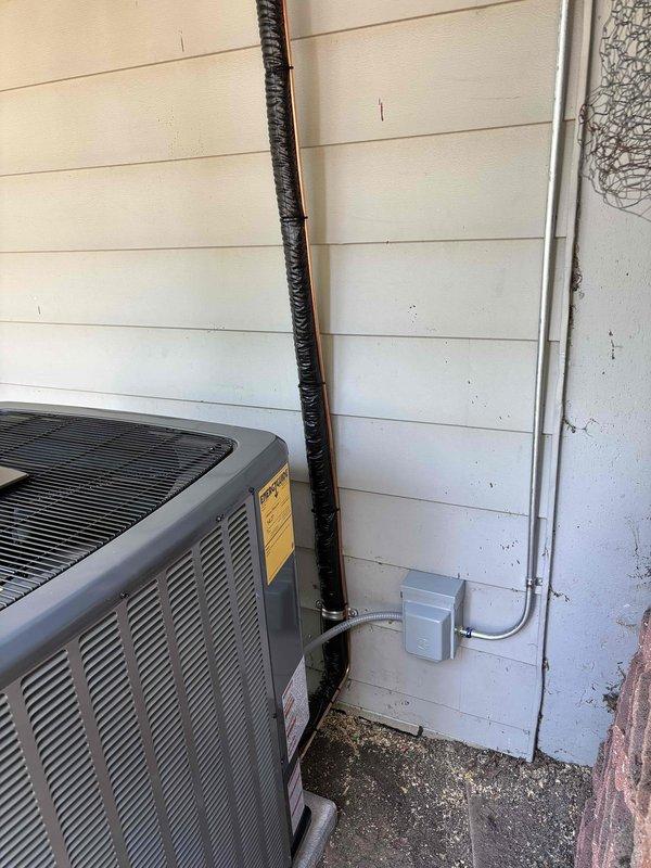 Completed add-on air conditioning system installation at residential property. Installed new gray condenser unit on concrete pad against exterior wall with proper refrigerant line set, electrical disconnect box, and metal conduit for power supply. All refrigerant lines properly insulated with black flexible insulation wrapping, system tested and operational.
