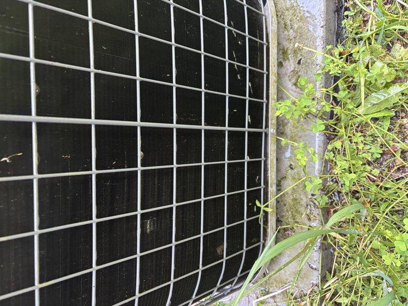 Responded to service call for indoor AC unit leaking water while still maintaining cooling capacity at single-family residence. Upon arrival, performed visual inspection of indoor air handler and outdoor condensing unit. Documented existing equipment conditions through photographs showing outdoor condenser unit positioned on concrete pad with vegetation growth around perimeter. Assessed drainage system components including condensate drain line, drain pan, and associated piping for blockages or improper pitch. Inspected evaporator coil for excessive dirt buildup or freezing conditions that could cause water overflow. Verified proper airflow across coil and checked air filter condition. Examined condensate pump operation if applicable and tested float switch functionality. Cleared any obstructions found in condensate drain line and verified proper water discharge. System continued operating with normal cooling function throughout service visit. Provided customer with findings and recomm