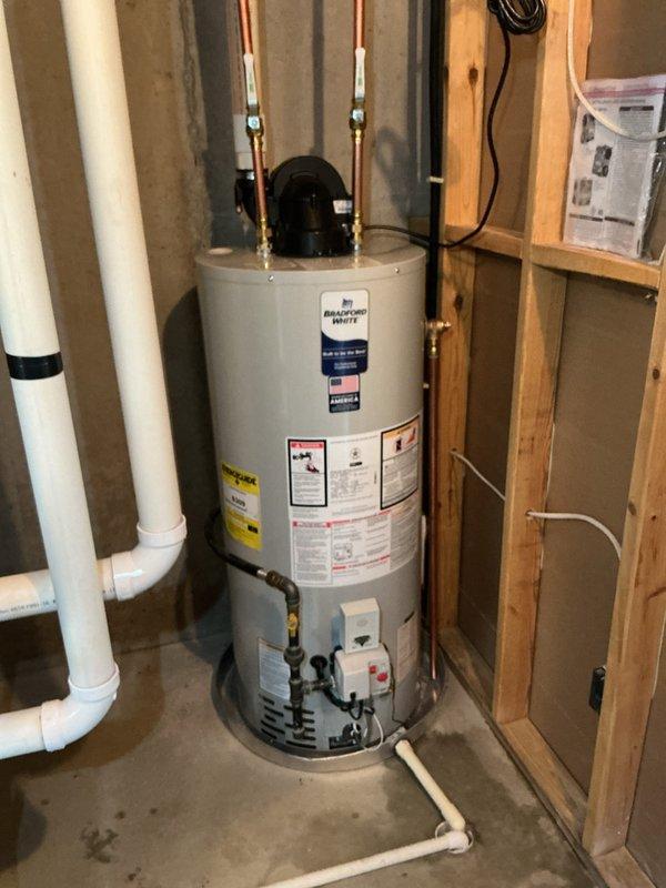 Completed installation of a 40-gallon Bradford White natural gas atmospheric vent water heater in residential basement utility space. Unit was properly positioned on concrete floor with copper supply lines and brass fittings connected at top of tank, circulation pump mounted at connections, and white insulation sleeves applied to exposed piping. All manufacturer specifications, safety labels, and operating instructions are clearly visible and accessible on unit exterior, with proper venting configuration established using existing PVC vent system.