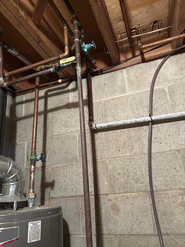 Responded to report of outside spigot leaking with visible water running and squirting from single location, noticed after sprinkler system activation. Upon arrival, inspected exterior hose bib and traced supply lines into basement utility area of 1972-built home. Located main water supply piping system with multiple copper lines showing age-related patina and corrosion. Identified shutoff valves serving exterior fixtures and documented existing plumbing configuration with aged copper supply lines, green-handled isolation valves, and various branch connections throughout exposed basement ceiling joists. Photographed plumbing infrastructure including insulated supply lines, flexible connections, and lockout-tagged equipment for service documentation. Customer reported concurrent issues with low pressure in certain sprinkler zones and non-functioning water softener, though stated belief that exterior leak was not irrigation-system related.