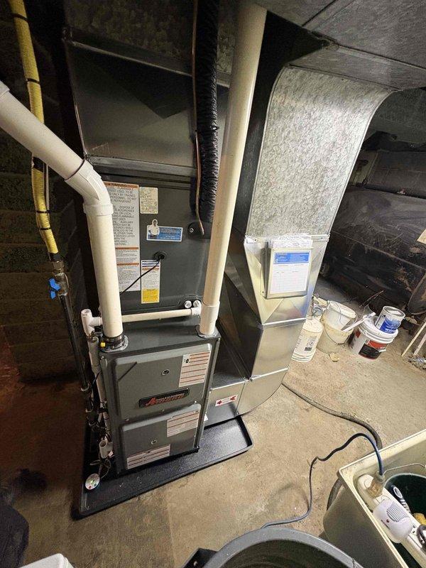 Completed full HVAC system installation including new Amana furnace unit and outdoor condensing unit. Furnace installed in basement utility area on protective mat with proper clearances, connected to evaporator coil unit with flexible ductwork, and integrated with existing gas line and PVC venting system routed through ceiling. Outdoor AC unit positioned on level concrete pad adjacent to exterior wall with refrigerant line sets and condensate drainage piping properly connected to interior equipment through wall penetration.