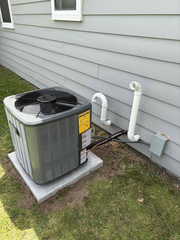 Completed full HVAC system installation including new Amana furnace unit and outdoor condensing unit. Furnace installed in basement utility area on protective mat with proper clearances, connected to evaporator coil unit with flexible ductwork, and integrated with existing gas line and PVC venting system routed through ceiling. Outdoor AC unit positioned on level concrete pad adjacent to exterior wall with refrigerant line sets and condensate drainage piping properly connected to interior equipment through wall penetration.