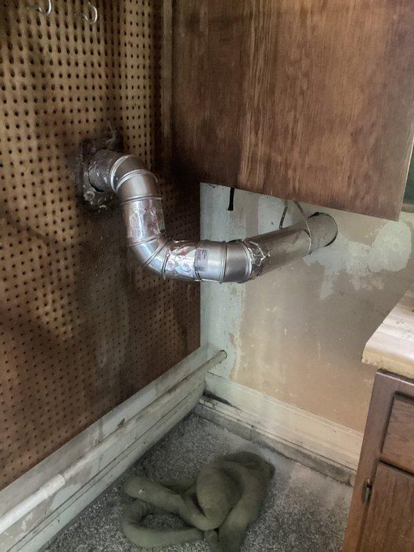# Dryer Vent Relocation to Exterior Wall

Successfully relocated dryer vent from existing configuration to exterior wall using flexible aluminum ductwork, routing through utility space with proper clearances and securing termination point through wall penetration. The previously scheduled faucet installation was cancelled per customer request, allowing focus on the ventilation project which was completed within the 6-hour timeframe.

## Work Performed

Assessed existing dryer vent configuration and determined optimal routing path to exterior wall. Created rectangular wall penetration in light-colored panel material to accommodate vent termination. Installed flexible aluminum ductwork with proper S-bend configuration to maintain airflow efficiency while navigating around existing pegboard and wood-paneled wall structures. Secured ductwork at mounting points and ensured proper connection at both appliance connection point and exterior wall termination. Verified ductwork was properly supp