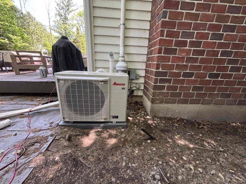 Completed full HVAC system installation including new furnace unit and Amana air conditioning condenser. Installed furnace in basement utility area with proper PVC venting routed vertically through structure and connected metallic plenum with galvanized ductwork for air distribution. Positioned outdoor AC condenser unit on concrete pad between brick wall and vinyl siding, ran refrigerant lines with white insulation wrapping, and connected to indoor system. All equipment secured, piping properly routed, and system tested for operational functionality.