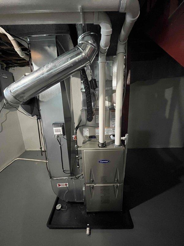 Completed installation of Carrier furnace unit in residential basement mechanical room. Unit properly mounted on black protective drip pan with all ductwork connections secured, including metallic supply plenum and curved trunk line extending to ceiling distribution system. Installed and secured white PVC condensate drain lines with appropriate mounting brackets and straps along vertical run. Verified all electrical connections, tested system operation, and confirmed proper airflow through supply ductwork. Installation meets manufacturer specifications and local code requirements.