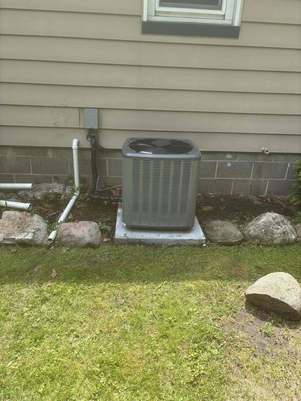 Completed full residential air conditioning system replacement including outdoor condensing unit and indoor furnace with integrated cooling coil. Installed new gray condensing unit on existing concrete pad against exterior wall with proper clearance and drainage considerations. Unit features horizontal ventilation slats and top-mounted fan housing. Connected white PVC condensate drainage lines along foundation. Indoor installation includes vertical furnace unit with gray metal cabinet mounted on black base platform in utility room. Integrated Aprilaire humidifier system to ductwork for whole-home humidity control. Installed white PVC venting system extending from furnace to exterior with proper pitch and support. Verified all electrical connections, refrigerant lines, and ductwork transitions between indoor and outdoor components. System tested and operational with all safety labels and compliance markings properly affixed to equipment.