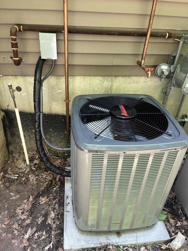 Completed full air conditioning system replacement at residential property. Removed existing outdoor condenser unit and installed new beige/tan cabinet condenser with black top-mounted fan assembly on existing concrete pad against building exterior. Reconnected copper refrigerant lines running vertically along wall, secured black insulated linesets, and verified all pipe connections for proper seal. Ensured electrical disconnect box remained accessible and properly mounted. Tested system operation, confirmed proper airflow through top-mounted fan grille, and verified all mechanical and electrical connections met manufacturer specifications. Unit installed on weathered concrete pad with all utility connections including gas meter clearances maintained per code requirements.