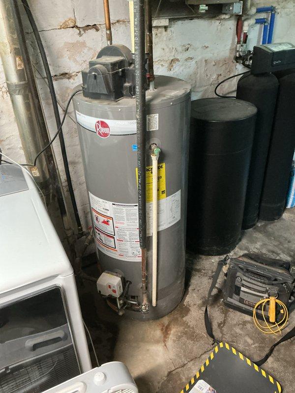 Completed comprehensive water heater estimate for existing Rheem unit located in basement utility area. Documented current installation configuration including all supply and discharge piping connections, electrical connections, temperature and pressure relief valve, and corrugated flex connectors at base. Photographed water heater condition showing age-related wear, surface staining, and accumulated dust on tank exterior. Assessed surrounding plumbing infrastructure including copper supply lines with visible oxidation and black flexible hoses. Noted presence of adjacent water treatment equipment including two black cylindrical filtration or softener tanks positioned near water heater. Evaluated overall mechanical room layout and accessibility for potential replacement work. Provided detailed estimate documentation with photographic evidence of existing conditions to customer for review and approval.