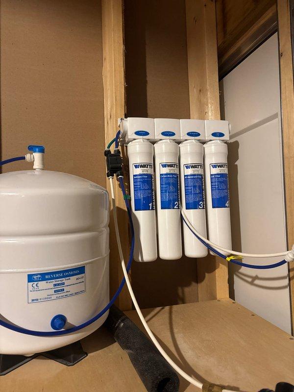 Responded to service call for malfunctioning reverse osmosis system. Upon inspection, documented the four-stage water filtration system installation including white pressure tank with blue valve assembly and vertically-mounted WATT brand filter cartridges numbered 1-4 connected via blue and beige tubing. System configuration and component layout were photographed for diagnostic purposes, with filtration assembly mounted to wall studs via black brackets and connected to pressure tank through manifold header. Customer approval obtained to proceed with troubleshooting and repair of the RO system malfunction.