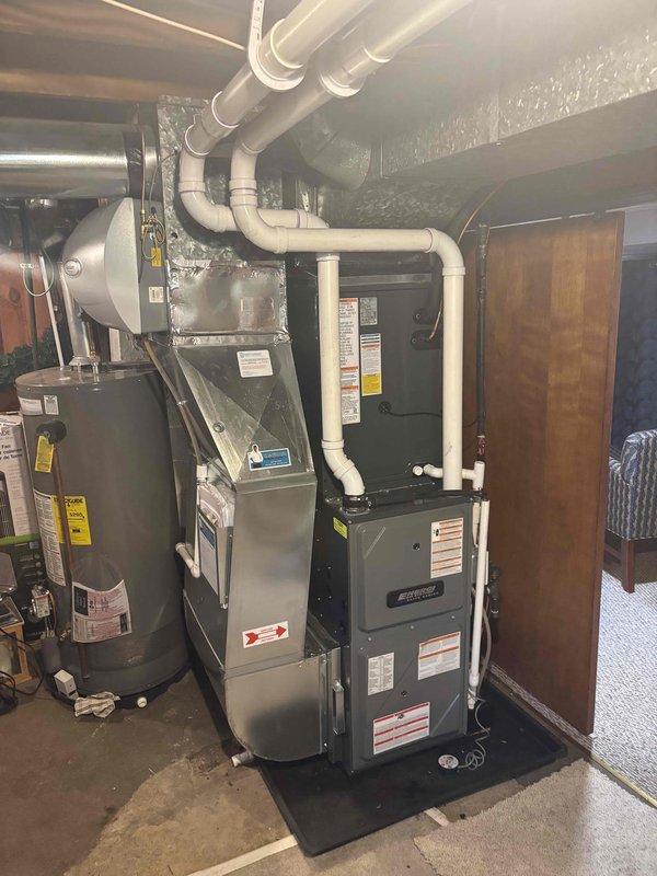 Completed full installation of new furnace, air conditioning system, and water heater in basement utility area. All units were properly positioned on protective mats over concrete flooring, with furnace equipped with white PVC venting routed overhead and connected to existing ductwork. Water heater installed adjacent to furnace with all necessary connections completed. All manufacturer labels and specifications visible on equipment, systems tested and verified operational upon completion.
