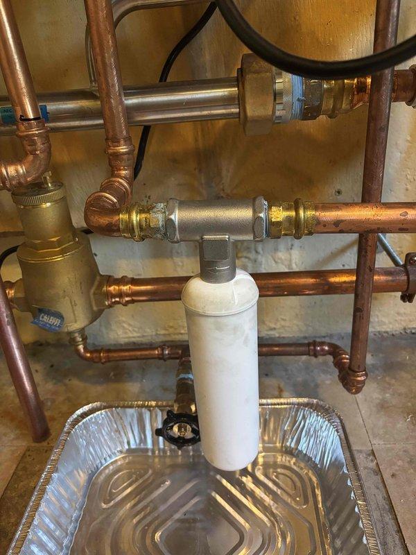 Responded to boiler leak complaint and inspected residential plumbing system. Upon arrival, identified active leak originating from severely corroded copper elbow joint with heavy oxidation and deterioration. Placed aluminum drip pans beneath affected area to contain water damage during assessment. Evaluated entire plumbing assembly including copper piping, brass fittings, compression joints, and pressure relief valve connections. Documented condition of corroded elbow joint and surrounding components through photographs. Per client request, relocated white cylindrical water filter to more accessible position within the plumbing configuration. Reconnected filter using appropriate brass compression fittings and verified secure connections throughout modified piping arrangement. System covered under home warranty program.