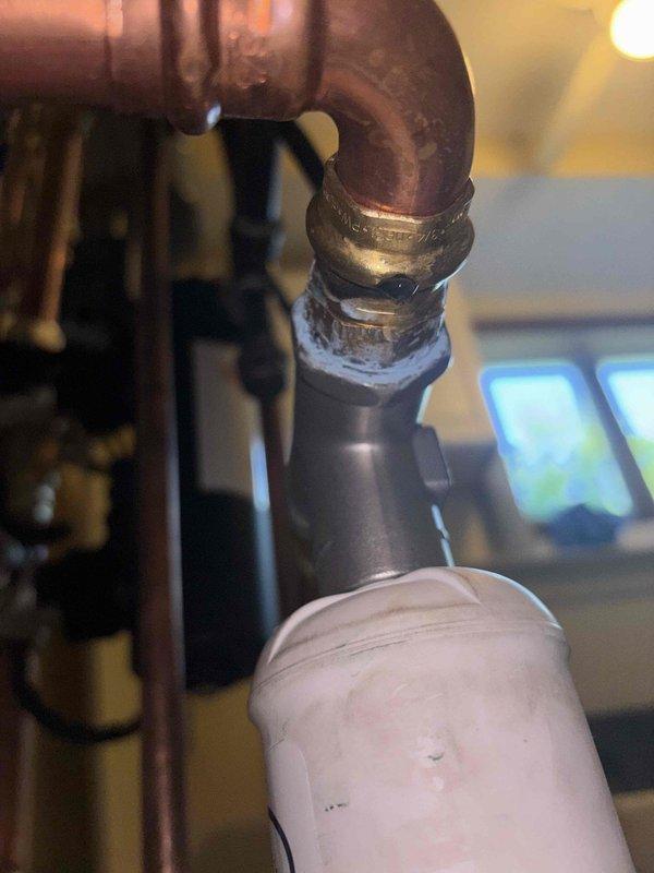Responded to boiler leak complaint and inspected residential plumbing system. Upon arrival, identified active leak originating from severely corroded copper elbow joint with heavy oxidation and deterioration. Placed aluminum drip pans beneath affected area to contain water damage during assessment. Evaluated entire plumbing assembly including copper piping, brass fittings, compression joints, and pressure relief valve connections. Documented condition of corroded elbow joint and surrounding components through photographs. Per client request, relocated white cylindrical water filter to more accessible position within the plumbing configuration. Reconnected filter using appropriate brass compression fittings and verified secure connections throughout modified piping arrangement. System covered under home warranty program.