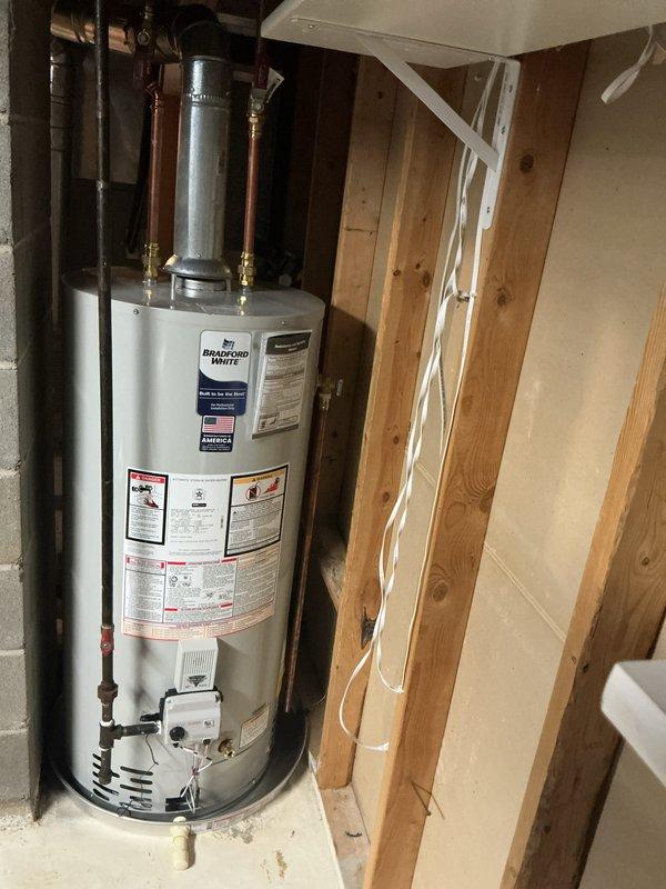 Completed installation of a 40-gallon Bradford White natural gas water heater with 6-year warranty in residential basement utility area. Unit installed as short model with proper venting, copper supply line connections with brass fittings, and black iron gas piping, positioned on concrete floor against exposed wall framing per manufacturer specifications and local building codes.