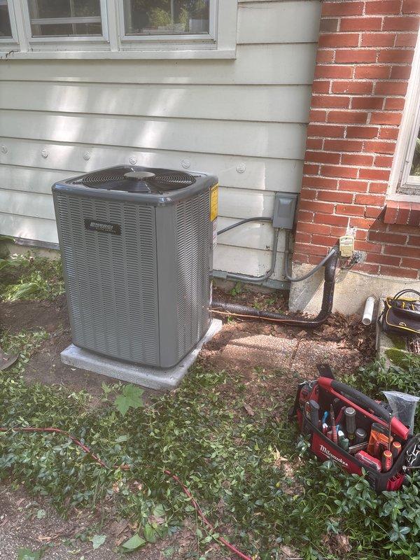 Complete air conditioning system replacement performed at residential property. Removed existing outdoor condenser unit and installed new gray/silver AC condenser on concrete pad between building siding and brick wall. Unit features mesh grille top with central fan hub and energy efficiency labeling. Properly secured refrigerant lines and electrical conduit along exterior wall using appropriate mounting hardware. Inside the basement utility area, coordinated the AC installation with existing Lennox furnace system and air handler/evaporator coil unit. Installed and configured white PVC condensate drainage piping with multiple 90-degree elbows, properly primed and secured to ceiling joists. Verified all electrical connections, refrigerant line sets, and control wiring between outdoor condenser and indoor components. System tested for proper operation, airflow, and temperature differential. Installation completed per manufacturer specifications with all safety labels and documentation aff
