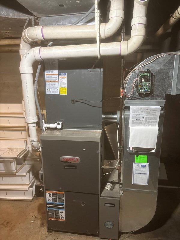 Complete air conditioning system replacement performed at residential property. Removed existing outdoor condenser unit and installed new gray/silver AC condenser on concrete pad between building siding and brick wall. Unit features mesh grille top with central fan hub and energy efficiency labeling. Properly secured refrigerant lines and electrical conduit along exterior wall using appropriate mounting hardware. Inside the basement utility area, coordinated the AC installation with existing Lennox furnace system and air handler/evaporator coil unit. Installed and configured white PVC condensate drainage piping with multiple 90-degree elbows, properly primed and secured to ceiling joists. Verified all electrical connections, refrigerant line sets, and control wiring between outdoor condenser and indoor components. System tested for proper operation, airflow, and temperature differential. Installation completed per manufacturer specifications with all safety labels and documentation aff