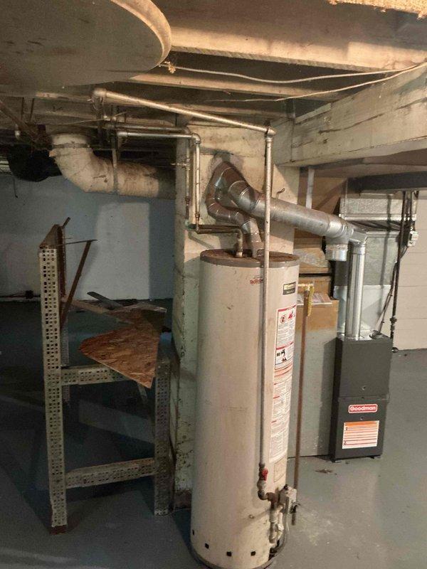Conducted on-site assessment of existing water heater in basement utility area for replacement estimate. Inspected aging cylindrical water heater unit showing significant corrosion and rust at base connections, particularly where copper supply lines interface with the tank. Documented condition of existing plumbing connections including copper and galvanized steel piping, vent stack configuration, and proximity to electrical panel. Evaluated basement mechanical room layout, ceiling clearances with exposed floor joists, and access requirements for equipment removal and installation. Photographed current water heater specifications, warning labels, and surrounding HVAC equipment to determine appropriate replacement unit sizing and compatibility with existing infrastructure.