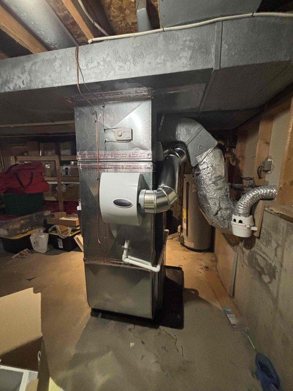 Completed full HVAC system installation including new furnace and air conditioning unit in basement mechanical room. Installed gray metal vertical furnace with integrated humidifier attachment featuring white cylindrical unit and metallic elbow connection. Configured flexible aluminum ductwork connections from furnace to existing rectangular duct system overhead. Installed air handler unit with copper refrigerant lines properly secured with metal straps along vertical run. Positioned equipment on black drain pan with white PVC condensate drain line for proper moisture management. Connected all ductwork, including flexible insulated ducting, to complete air distribution system. Verified all mechanical connections, electrical hookups, and system integration. System tested and operational upon completion.