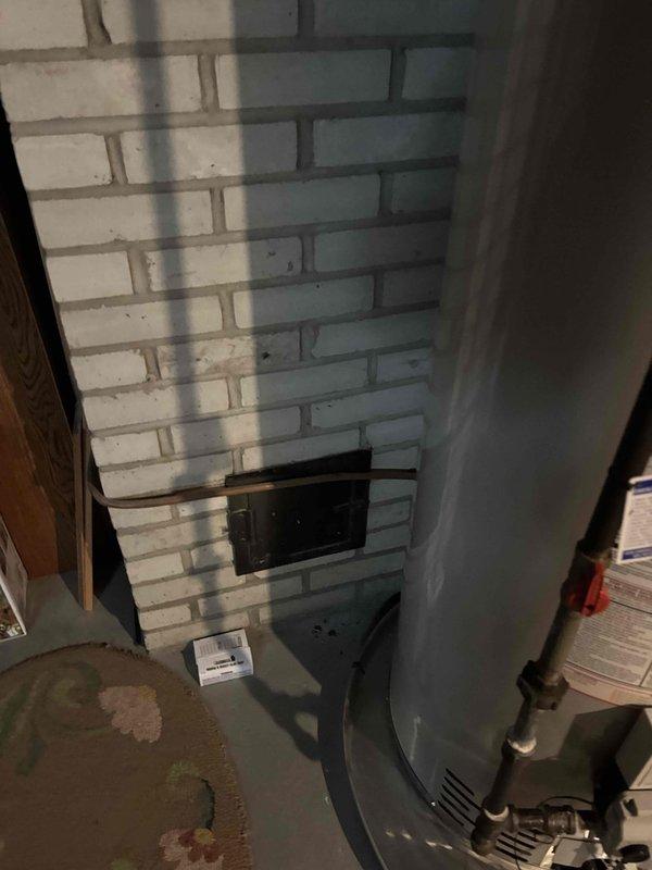 Responded to call regarding boiler making unusual noise while thermostat was in off position during warm weather. Upon arrival, inspected 22-year-old boiler system and found evidence of previous combustion issues indicated by sooting and dark staining on white brick chimney wall surrounding flue access panel. Examined boiler operation, venting system, and flue connections. Documented existing conditions including discoloration on masonry and floor staining in utility area. Advised customer of system age and observations regarding combustion residue patterns on surrounding surfaces.