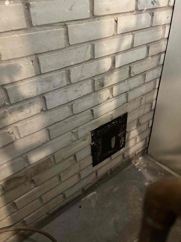 Responded to call regarding boiler making unusual noise while thermostat was in off position during warm weather. Upon arrival, inspected 22-year-old boiler system and found evidence of previous combustion issues indicated by sooting and dark staining on white brick chimney wall surrounding flue access panel. Examined boiler operation, venting system, and flue connections. Documented existing conditions including discoloration on masonry and floor staining in utility area. Advised customer of system age and observations regarding combustion residue patterns on surrounding surfaces.