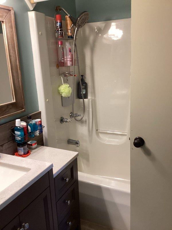 Conducted on-site assessment of existing bathtub-shower combination unit for conversion to walk-in shower installation. Documented current bathroom configuration including white fiberglass tub surround, chrome fixtures, grab bar placement, and vanity positioning. Photographed existing plumbing connections, shower valve assembly, and tub drain location to prepare specifications for tub removal and barrier-free shower replacement.