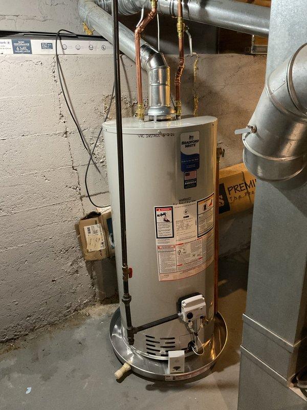 Completed installation of a 40-gallon natural gas water heater in residential basement utility room. Unit was installed on proper drip pan with all copper supply lines, gas connections, and exhaust venting properly configured and secured. Bradford White water heater was positioned with appropriate clearances, all plumbing connections were completed with brass fittings, and flue pipe was connected to existing HVAC ductwork system. Installation includes proper electrical connections and temperature controls, with all manufacturer specifications and local code requirements met for safe operation.