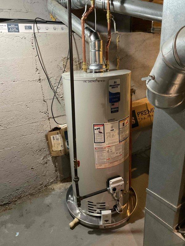 Completed installation of a 40-gallon natural gas water heater in residential basement utility room. Unit was installed on proper drip pan with all copper supply lines, gas connections, and exhaust venting properly configured and secured. Bradford White water heater was positioned with appropriate clearances, all plumbing connections were completed with brass fittings, and flue pipe was connected to existing HVAC ductwork system. Installation includes proper electrical connections and temperature controls, with all manufacturer specifications and local code requirements met for safe operation.