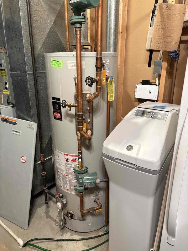 Conducted estimate for water heater replacement in residential utility room. Evaluated existing tall white cylindrical water heater with copper piping connections, brass valves, and circulation pump mounted at base. Assessed current plumbing configuration including multiple shut-off valves with blue handles and complex copper pipe routing. Documented equipment layout in unfinished basement space alongside existing HVAC furnace and water softener system. Photographed installation conditions and provided customer with replacement options and recommendations for new water heater installation.