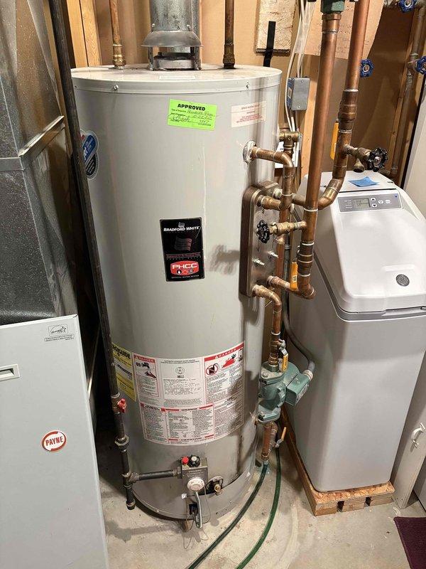 Conducted estimate for water heater replacement in residential utility room. Evaluated existing tall white cylindrical water heater with copper piping connections, brass valves, and circulation pump mounted at base. Assessed current plumbing configuration including multiple shut-off valves with blue handles and complex copper pipe routing. Documented equipment layout in unfinished basement space alongside existing HVAC furnace and water softener system. Photographed installation conditions and provided customer with replacement options and recommendations for new water heater installation.