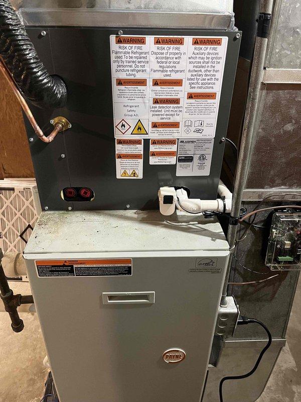 Completed full HVAC system replacement including outdoor condensing unit and indoor air handler with furnace. Installed new dark gray condenser on existing concrete pad with proper electrical disconnect, refrigerant lines, and drainage connections. Mounted new air handler above existing furnace in basement mechanical room, connected copper refrigerant lines, PVC condensate drain, and integrated with existing ductwork using flexible transitions. System tested and verified operational with all safety labels applied per manufacturer specifications.