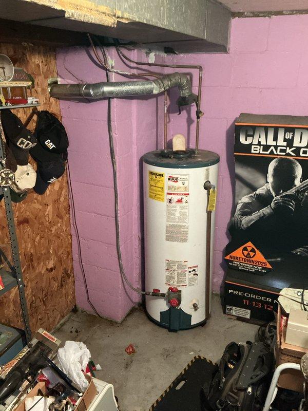 Responded to water heater leak from top area on 5-year-old unit. Upon inspection, identified moisture accumulation on pipes and connections above RUUD water heater in basement utility area. Assessed plumbing connections, copper piping network, and condensation patterns on metallic ductwork and pipes. Documented existing installation configuration including water heater placement, pipe routing, and connection points for diagnostic purposes.