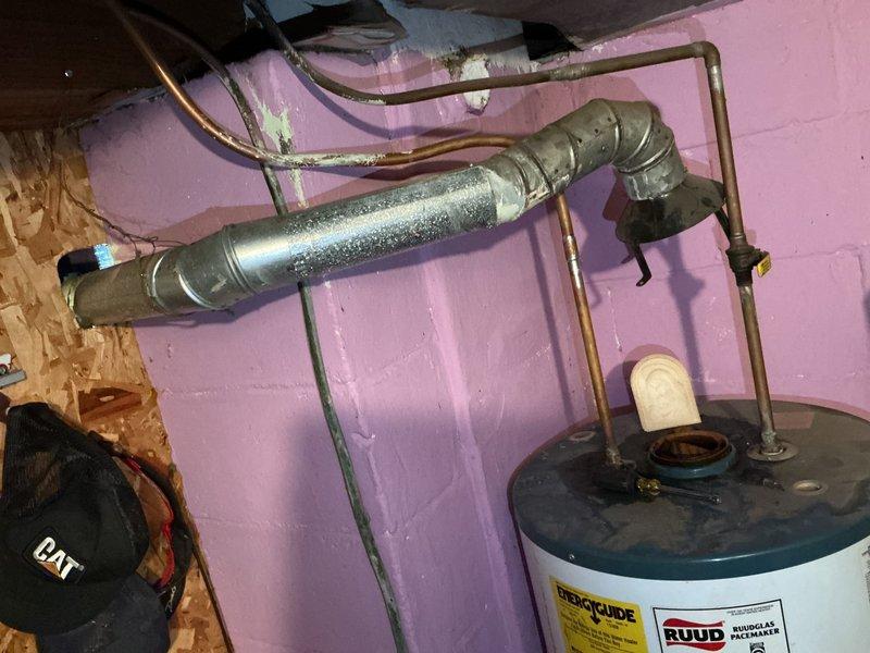 Responded to water heater leak from top area on 5-year-old unit. Upon inspection, identified moisture accumulation on pipes and connections above RUUD water heater in basement utility area. Assessed plumbing connections, copper piping network, and condensation patterns on metallic ductwork and pipes. Documented existing installation configuration including water heater placement, pipe routing, and connection points for diagnostic purposes.