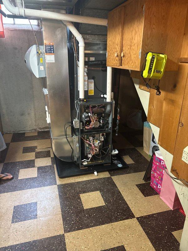 Completed full HVAC system installation including new furnace and air conditioning unit. Interior furnace installed in basement utility area with all internal components properly connected, including copper refrigerant lines, electrical wiring, and motor assemblies integrated with existing ductwork system. Exterior condenser unit positioned on concrete pad against building exterior with refrigerant lines and electrical connections routed through wall penetration, condensate drain line secured along exterior wall to electrical disconnect box. Both units tested and verified operational upon completion of installation.