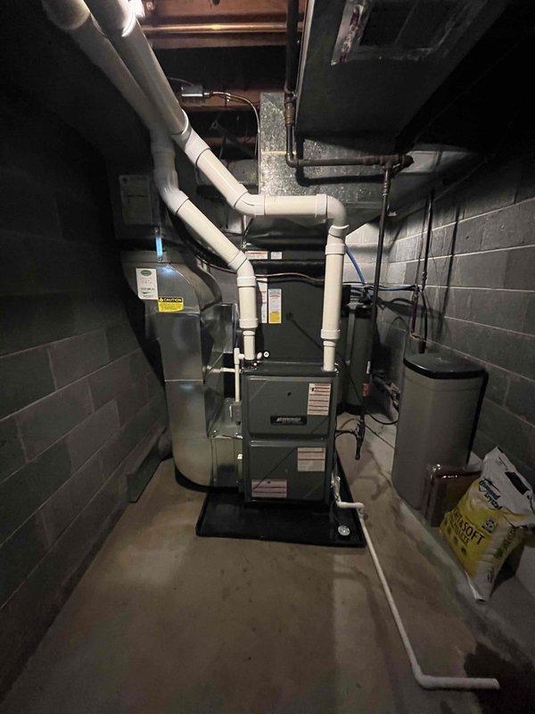 Completed full HVAC system installation including new furnace and air conditioning unit. Interior furnace installed in basement mechanical room with proper venting through white PVC exhaust and intake piping, electrical connections established, and all safety labels verified. Exterior condensing unit positioned on concrete pad with decorative rock surround, refrigerant lines connected and insulated, service valves installed with protective caps, and unit fully operational and tested for proper function.