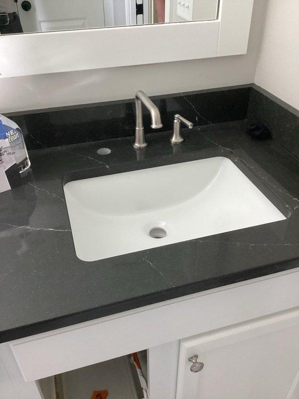 Completed plumbing project estimate for bathroom vanity reinstallation following new countertop and sink installation. Assessed two vanity locations: first bathroom features dark charcoal countertop with white veining and undermount white sink with brushed nickel three-piece faucet set; second bathroom has white veined countertop with undermount rectangular sink and matte black dual-handle centerset faucet. Documented existing conditions including visible surface staining on white countertop requiring attention, confirmed all plumbing connections accessible through white cabinetry, and verified faucet mounting configurations for proper reinstallation procedures.